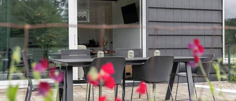 Un patio exterior con mesa y sillas, un ventanal de vidrio que da a una habitación y vistas a la vegetación.