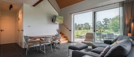 A modern living room with a sofa, a coffee table, and a dining area with chairs and a table. There is a staircase leading to another level and a large window with a view of greenery outside.