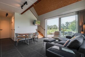 A modern living room with a sofa, a coffee table, and a dining area with chairs and a table. There is a staircase leading to another level and a large window with a view of greenery outside.