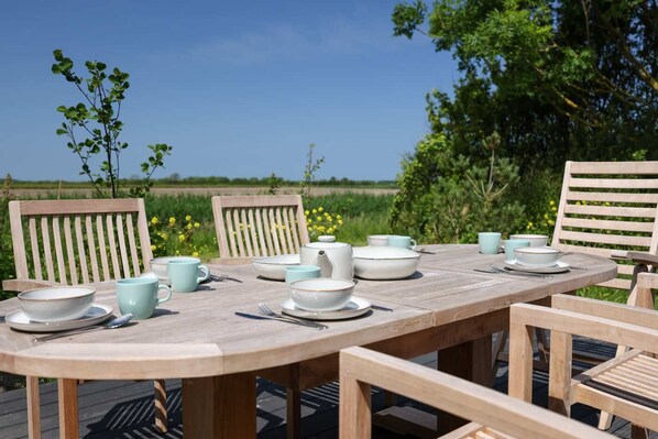 Une table en bois, dressée pour un repas en plein air, avec des chaises, des bols, des tasses et une théière.