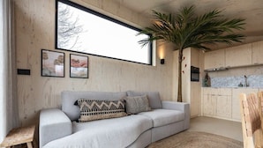 A modern living room with a curved grey sofa, a large window, and a kitchen area with wooden cabinets.