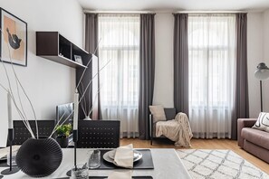 A modern living room with a sofa, a dining table set, and a chair. There are two large windows with curtains, a black bookshelf, and a floor lamp. - Black Swan Apartment - 50 Meters from Dancing House (Hlavní město Praha)