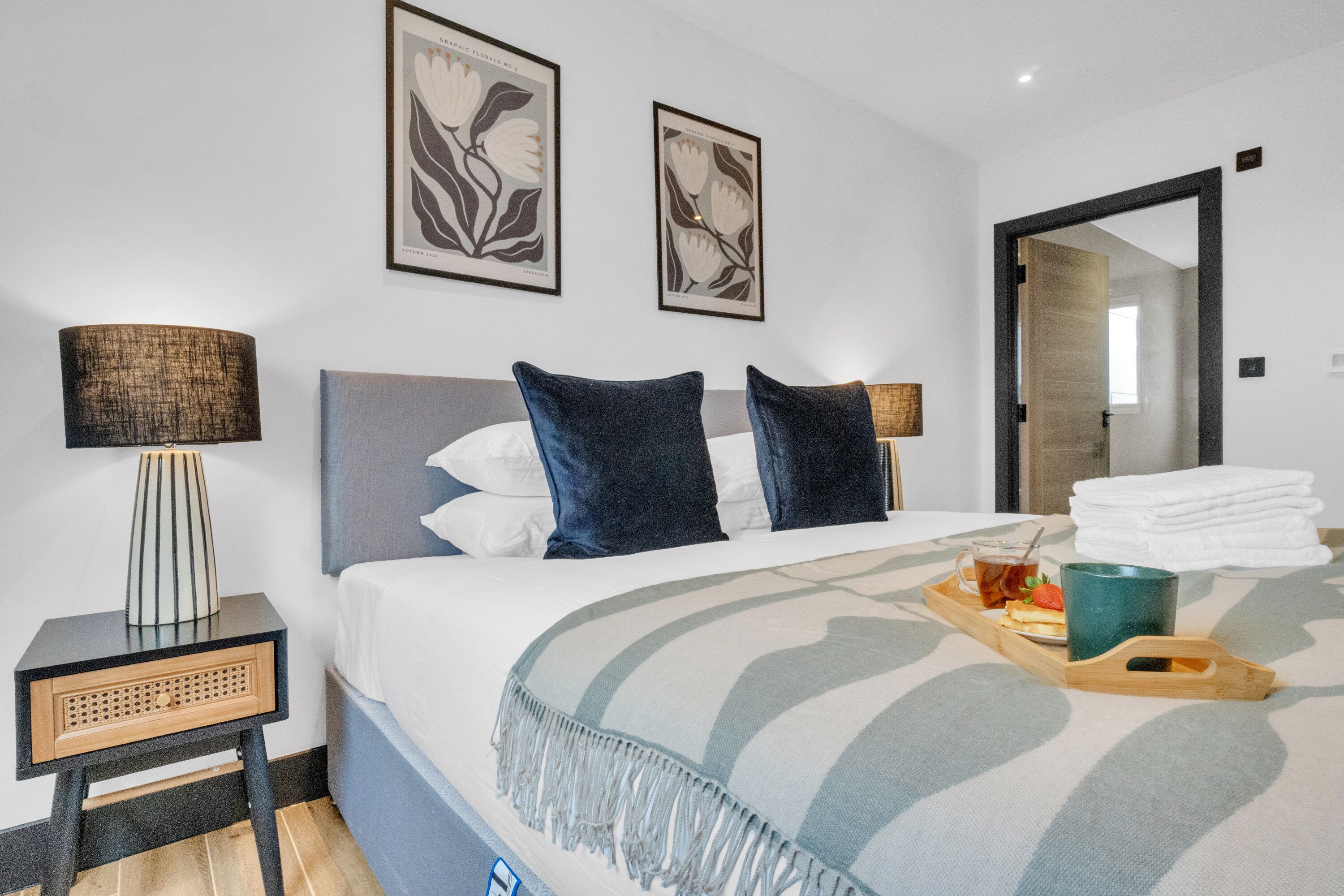 A neatly made bed with a striped blanket, two pillows, and a tray with a cup and a plate of food. There are two wall-mounted lamps and framed artwork above the bed.