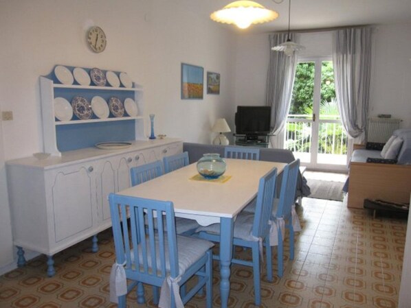 Un coin repas avec une table blanche et des chaises bleues, une cuisine avec des meubles blancs, un téléviseur et un balcon avec des rideaux.