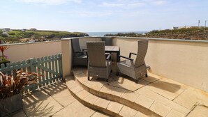 A patio with wicker chairs and a table, overlooking a coastal landscape with houses and greenery. - Beachside apartment with a private patio garden and beautiful sea views (Padstow)