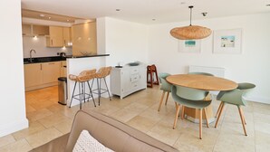 A modern kitchen with a dining area, featuring a round wooden table, green chairs, and a woven pendant light. - Beachside apartment with a private patio garden and beautiful sea views (Padstow)