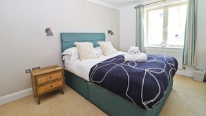 A neatly made bed with a teal headboard, white bedding, and a dark blue blanket. There is a small wooden nightstand beside the bed with a lamp on it. A window with curtains is visible in the background. - Beachside apartment with a private patio garden and beautiful sea views (Padstow)