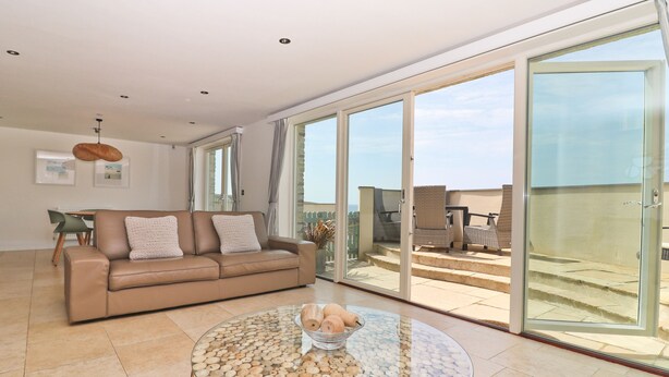A modern living room with a glass coffee table, a beige sofa, and a large sliding glass door leading to a balcony. - Beachside apartment with a private patio garden and beautiful sea views (Padstow)