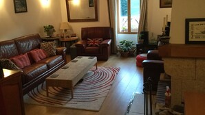 Ein gemütliches Wohnzimmer mit einem Ledersofa, einem Couchtisch aus Holz und einem roten Teppich. An der Wand hängt ein großer Spiegel, ein Fenster mit Vorhängen und ein Kamin mit Kaminfeuerbesteck.