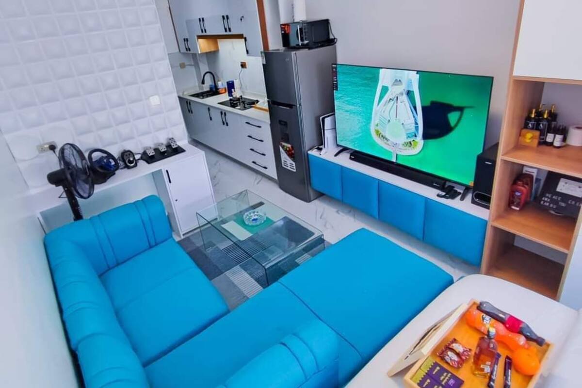 Un salon moderne avec un canapé bleu, une table basse en verre, un téléviseur à écran plat et un coin cuisine doté de meubles blancs et d’un four à micro-ondes.