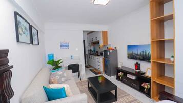 Una sala de estar moderna con un sofá, una mesa de centro y un televisor. Se aprecia una cocina al fondo, con armarios y un refrigerador.