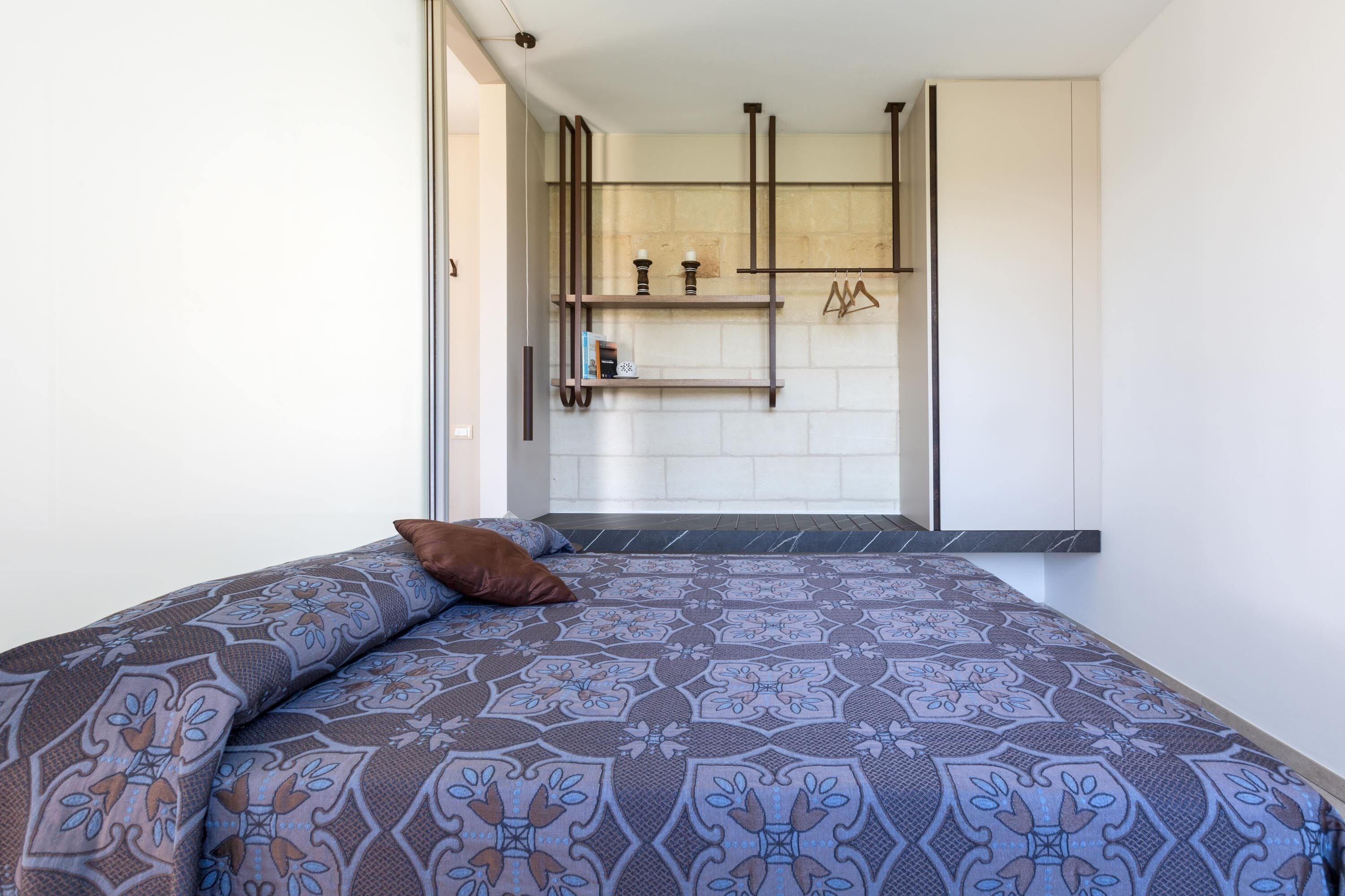 Een slaapkamer met een bed met een patroon, een bruin kussen en een ingebouwde plank met decoratieve objecten.