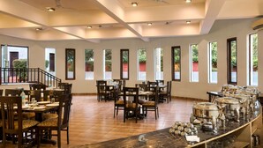 A spacious dining area with multiple tables and chairs, large windows with views of greenery, and ceiling fans. - Hridey Retreat Resort (Ramnagar)