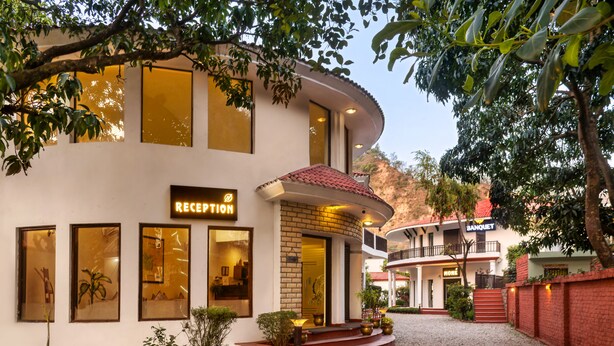 A reception area with a sign, a curved building facade, and a gravel driveway leading to a building with a red roof. - Hridey Retreat Resort (Ramnagar)