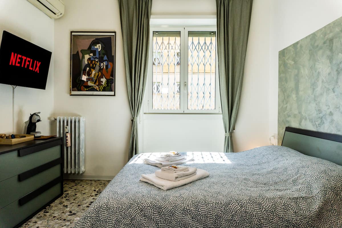 A bedroom with a bed, a dresser, a radiator, a window with curtains, a painting, and a TV displaying 'NETFLIX'.
