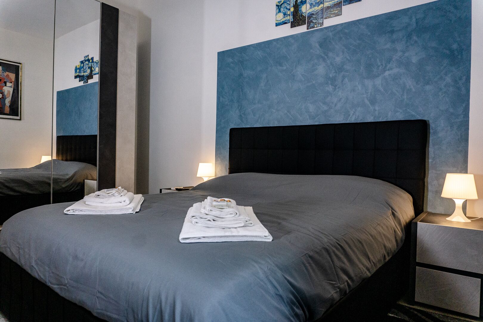 A hotel room with a large bed, two bedside tables, a nightstand, a mirror, and artwork on the wall.