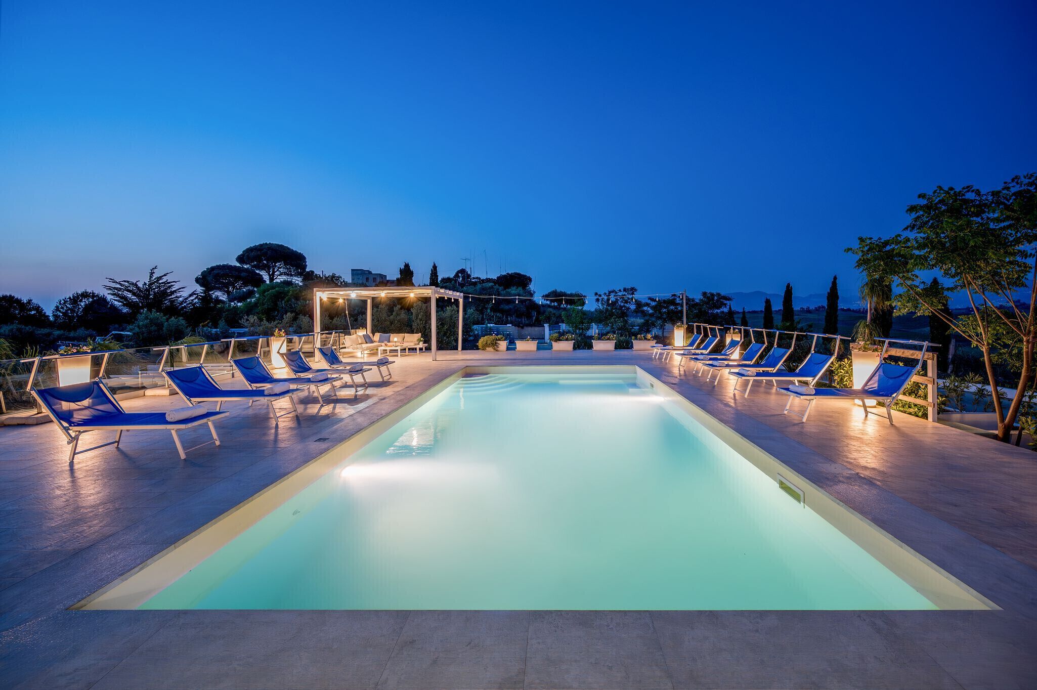 Una piscina con sillas de descanso y una pérgola, rodeada de árboles y edificios.