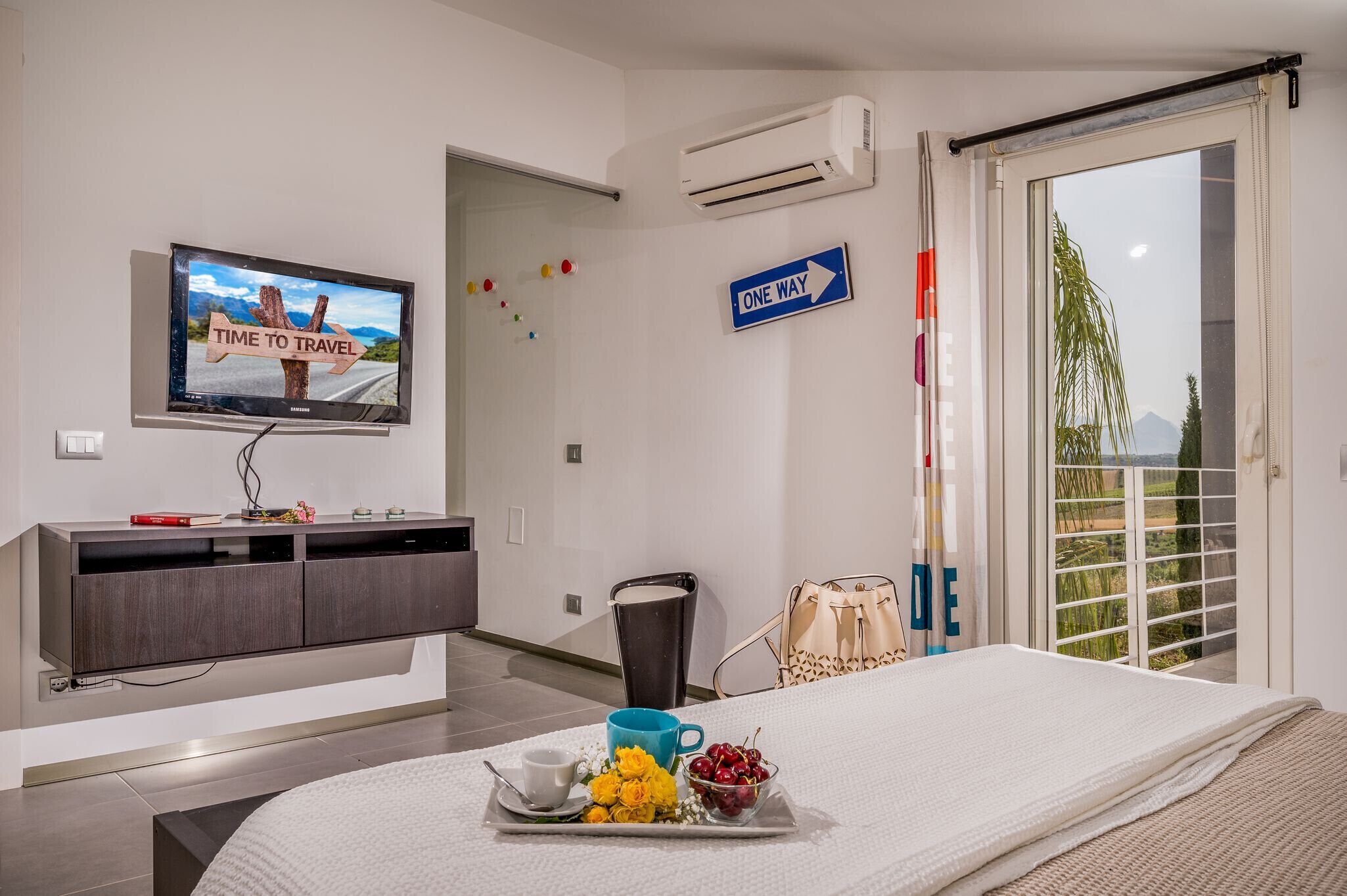 Une chambre d’hôtel avec un lit, un téléviseur affichant une image sur le thème du voyage, un panneau indiquant « SENS UNIQUE » et une vue sur la verdure par la fenêtre.