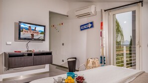 Ein Hotelzimmer mit Bett, Fernseher der ein Reisebild zeigt, einem 'ONE WAY'-Schild und Blick auf Grün durch das Fenster.