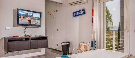 A hotel room with a bed, a TV displaying a travel-themed image, a 'ONE WAY' sign, and a view of greenery through the window.