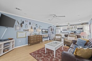A living room with a TV, wooden furniture, a ceiling fan, and a kitchen in the background.