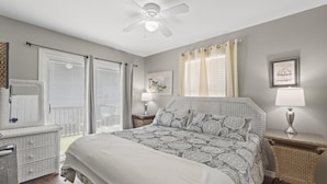 Un dormitorio con cama, ventilador de techo, mesita de noche con lámpara, cómoda y ventana con cortinas.