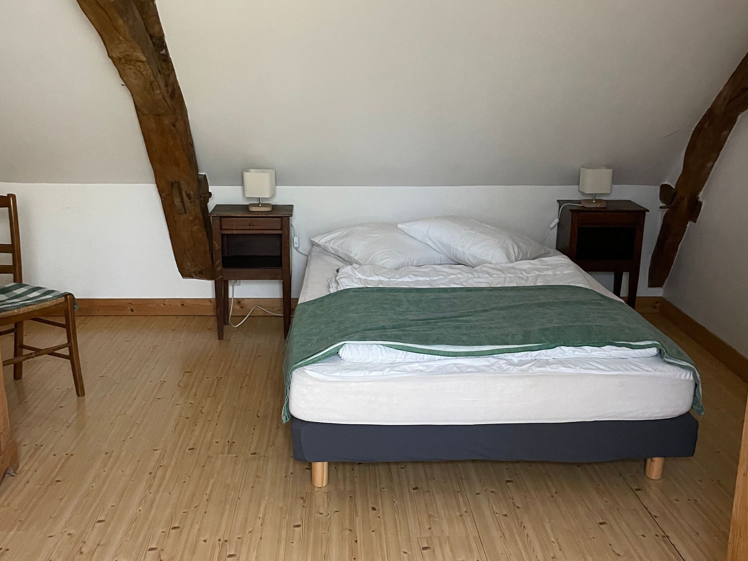 A bedroom with a bed, two bedside tables, a chair, and wooden beams on the ceiling.