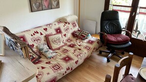 A cozy living room with a patterned sofa, a black office chair, a wooden dining table, and a wall clock. - Charming T3 in Combloux Center - Pets and Parking Included (Combloux)