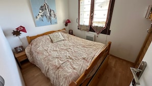 A bedroom with a wooden bed, a painting of a mountain, a window with curtains, and a bedside lamp. - Charming T3 in Combloux Center - Pets and Parking Included (Combloux)