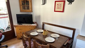 A dining area with a wooden table set for four, a television above the table, and a cabinet with glass doors. - Charming T3 in Combloux Center - Pets and Parking Included (Combloux)