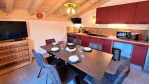 Una cocina moderna con armarios rojos, una zona de comedor con una mesa grande dispuesta para seis personas y un televisor sobre un soporte de madera.
