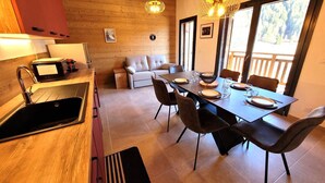 Una cocina moderna con un lavamanos, estufa y microondas. Hay una zona de comedor con una mesa dispuesta para cuatro, sillas y un sofá. La habitación tiene paredes de madera y ventanas amplias.