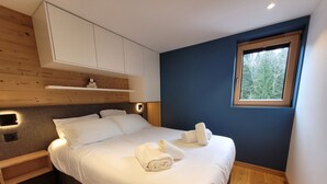 Ein modernes Schlafzimmer mit Bett, weißen Bettwäsche, einem Holz-Kopfteil, Einbauschränken, einer kleinen Ablagefläche, einem Nachttisch, einer Wandleuchte und einem Fenster mit Blick auf Bäume.