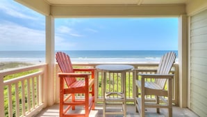 Ein überdachter Balkon mit zwei Adirondack-Stühlen und einem kleinen Tisch, der auf einen Strand und das Meer blickt.