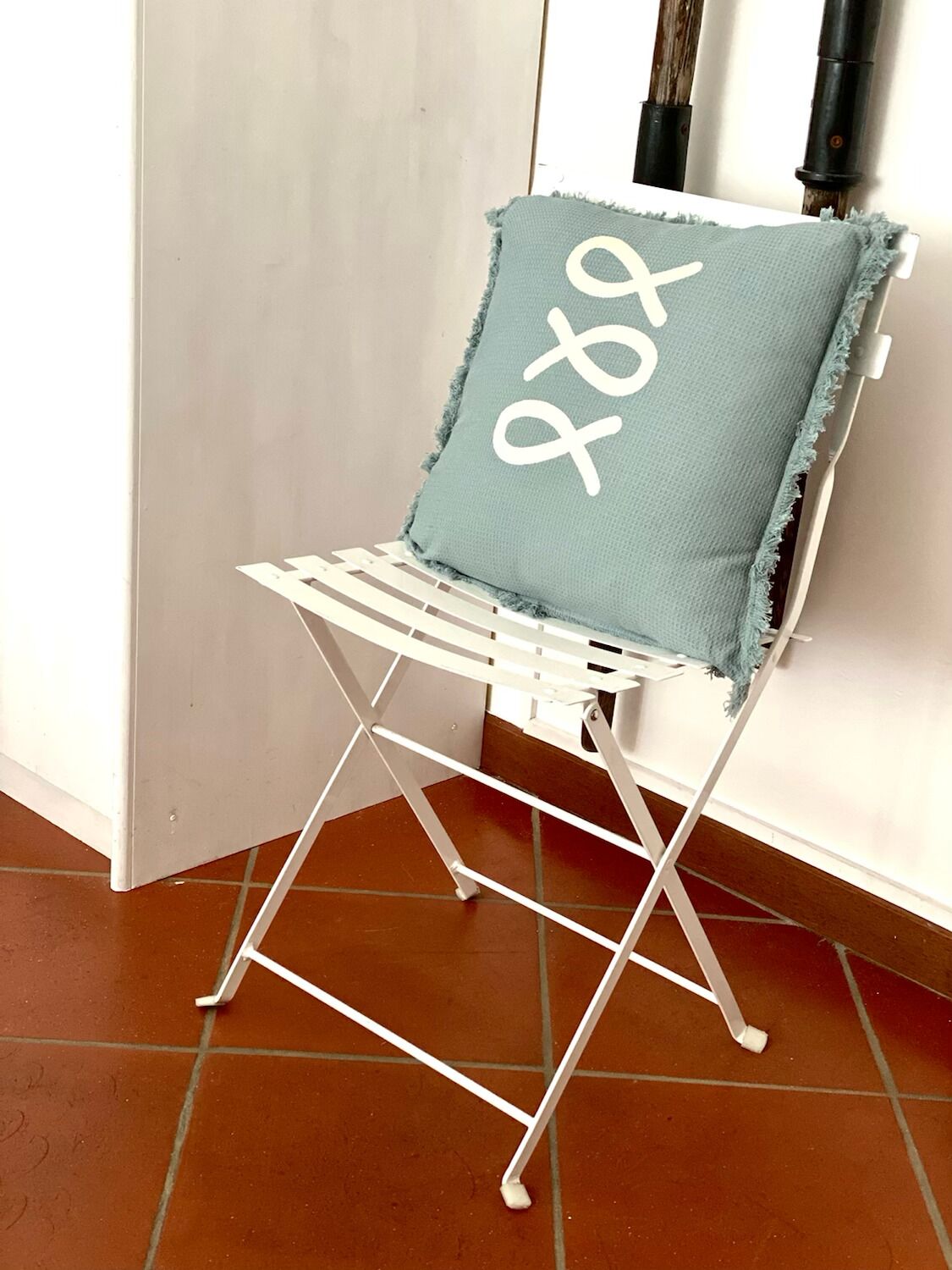 A white folding chair with a light blue cushion featuring a white anchor design, positioned against a white wall with a tiled floor.