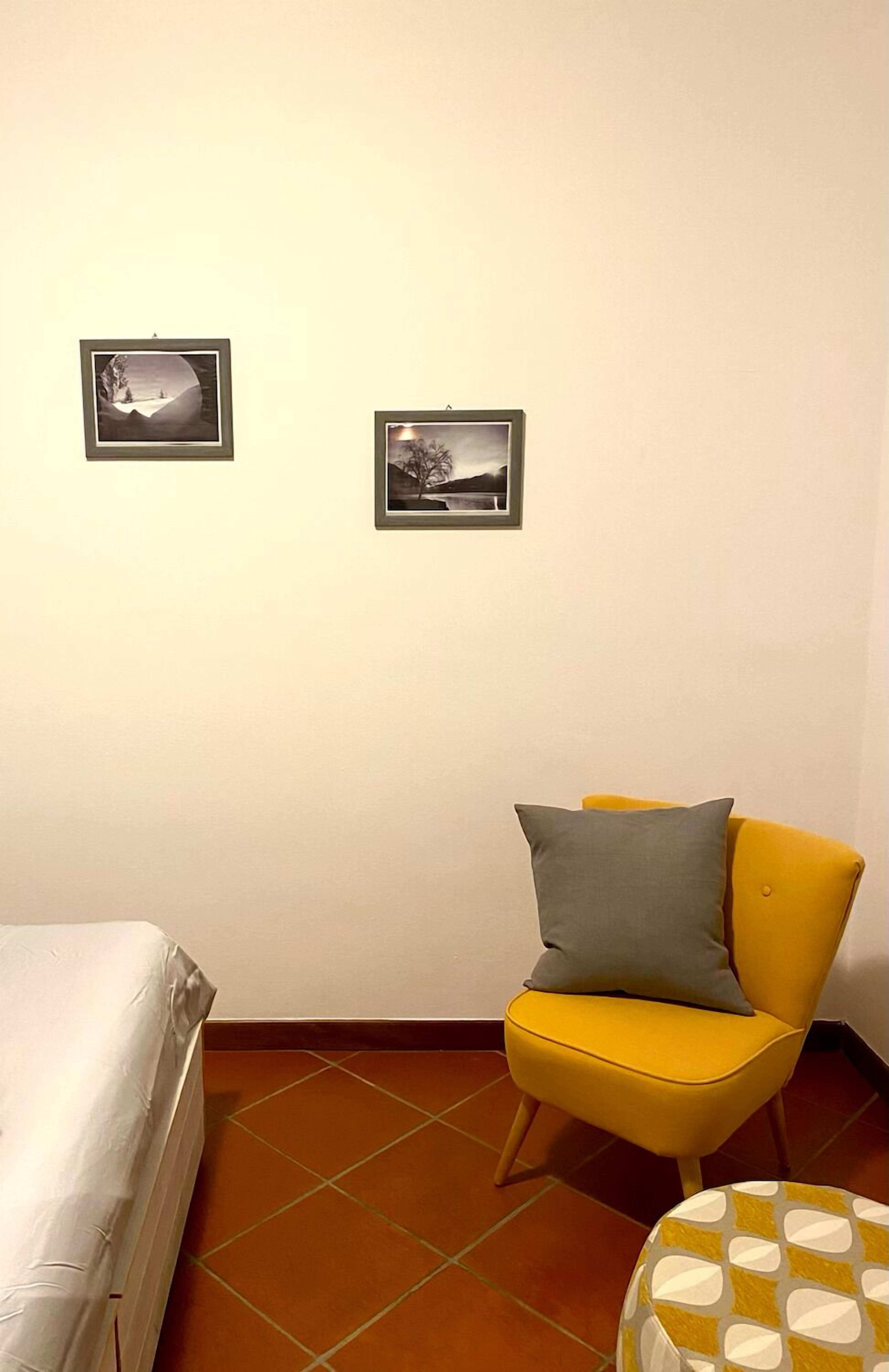 Una stanza con un letto, una sedia gialla con un cuscino e due quadri appesi al muro.