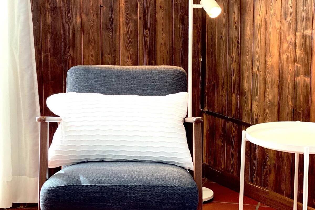 Une chaise avec un coussin bleu et un oreiller blanc, adossée à un mur en bois, une table blanche et un lampadaire.