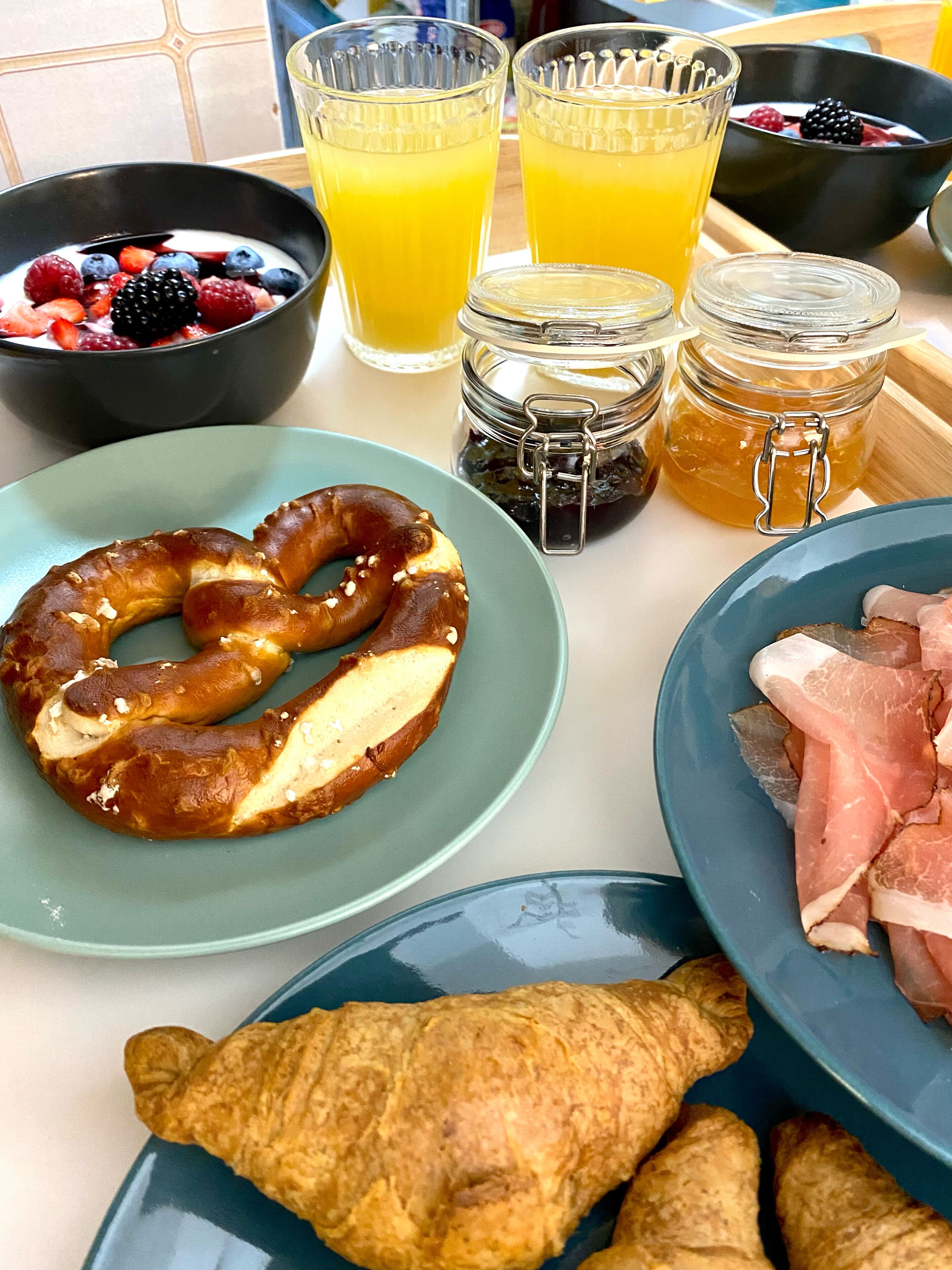 A breakfast table with a pretzel, croissant, sliced ham, a bowl of berries, and a glass of orange juice.