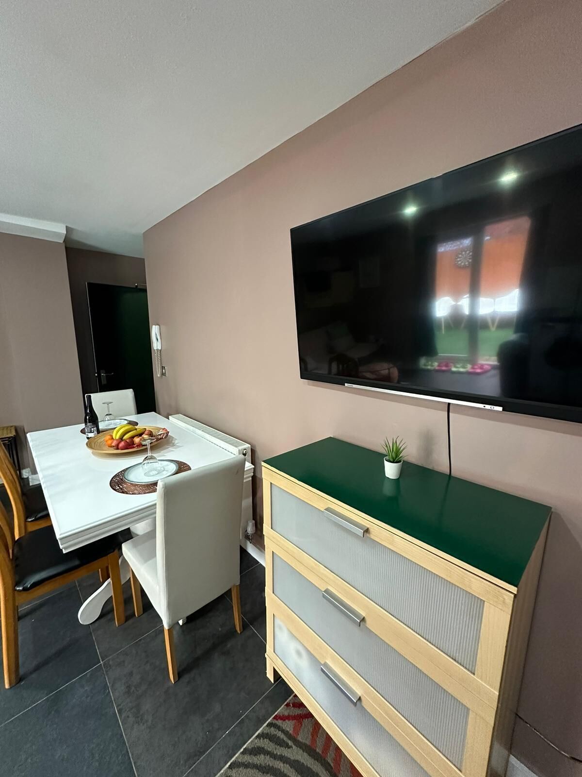 A modern living room with a flat-screen TV mounted on the wall, a dining table set with fruits and a wine glass, and a green cabinet with drawers.