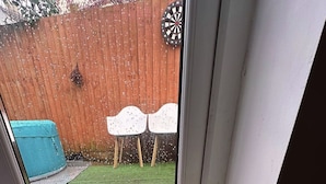 Une fenêtre donnant sur un jardin, avec une clôture en bois, un jeu de fléchettes et deux chaises blanches.