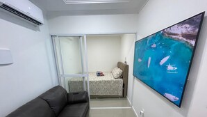 Quarto de hotel moderno com sofá de couro preto, cama com colcha floral, televisão de tela plana grande na parede e porta de vidro deslizante que dá para uma varanda.