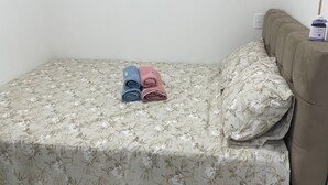Cama pequena e bem arrumada, com colcha florida e duas toalhas enroladas em azul e rosa.