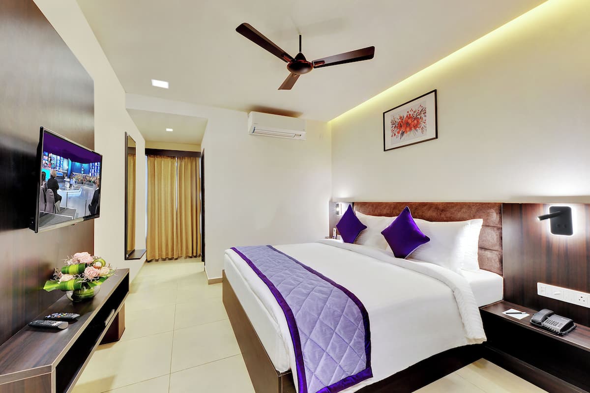 A hotel room with a large bed, a flat-screen TV, a ceiling fan, an air conditioning unit, and a bedside table with a phone and remote controls.