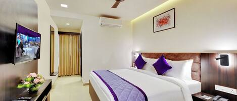 A hotel room with a large bed, a flat-screen TV, a ceiling fan, an air conditioning unit, and a bedside table with a phone and remote controls.