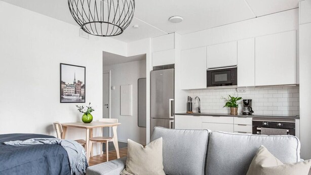 Eine moderne Wohnung mit grauem Sofa, einem Esstisch mit Stühlen, einer Küche mit weißen Schränken und einer Pendelleuchte.
