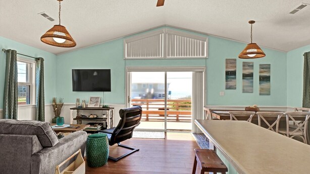 Uma sala de estar espaçosa com teto alto, área de jantar com uma mesa comprida e vista para a praia através de uma porta de vidro deslizante.