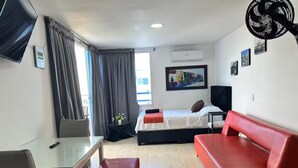 Una habitación de hotel con una cama, un sofá rojo, un escritorio con una silla, un televisor y un equipo de aire acondicionado.
