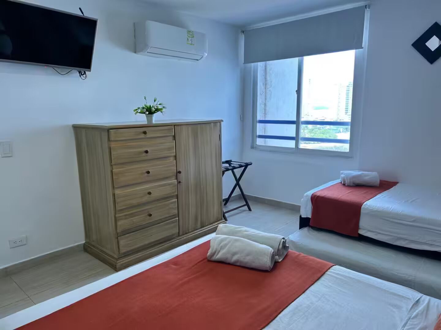 A hotel room with two beds, a TV, an air conditioner, a window with a view, and a small table.