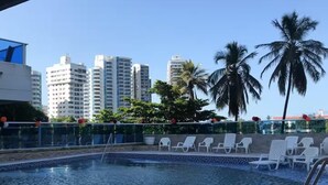 Una piscina con sillas de descanso, palmeras y edificios altos al fondo.