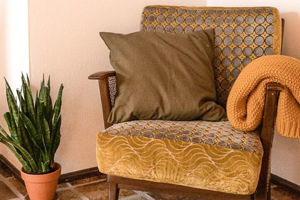 Een hoek van een kamer met een houten stoel, een gekleurd kussen, een plant in pot en een gebreide plaid.